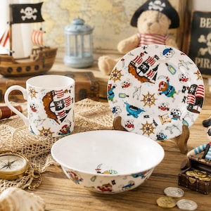 May include: A white ceramic dinnerware set consisting of a mug, a bowl, and a plate, all decorated with a colourful pirate-themed pattern featuring ships, treasure maps, and cartoon characters on a white background.