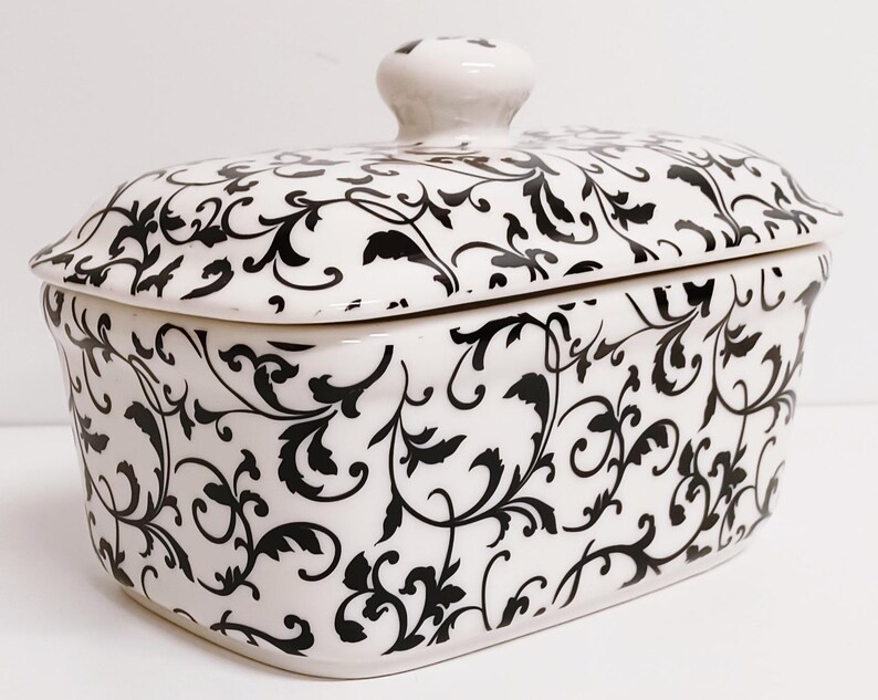 K&ouml;nnte beinhalten: Eine wei&szlig;e Keramik-Butterdose mit einem schwarz-wei&szlig;en Blumenmuster. Der Deckel hat einen Knopf oben.