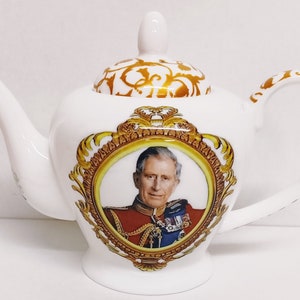 Op de afbeelding: Witte porseleinen theepot met een gouden rand en een portret van koning Charles III in een gouden lijst. De deksel van de theepot heeft een gouden bloemmotief.