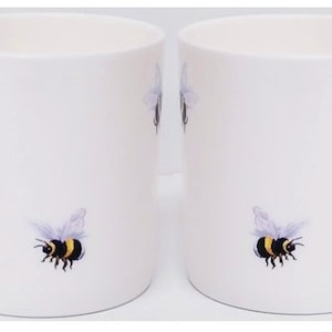 May include: Two white ceramic mugs with black and yellow bee illustrations. Each mug has three bees, two on the front and one on the side.