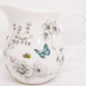 May include: White ceramic creamer with a floral and butterfly design. The design features gray flowers, green leaves, a blue butterfly, and a yellow and black bee.