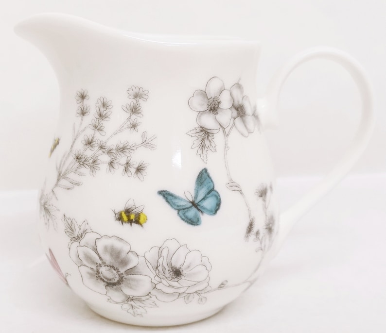 May include: White ceramic creamer with a floral and butterfly design. The design features gray flowers, green leaves, a blue butterfly, and a yellow and black bee.