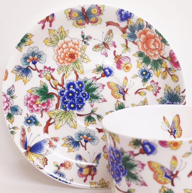 May include: White teacup and saucer set with a colorful floral and butterfly design. The pattern includes blue, pink, orange, and yellow flowers and butterflies. The teacup and saucer are likely made of porcelain.