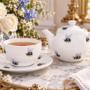 May include: A white ceramic teapot and a matching teacup with a saucer, both decorated with a repeating pattern of small bumblebees with yellow and black bodies and light blue wings. The teapot features a lid with a matching bee illustration.