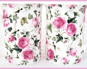May include: Two white ceramic mugs with pink rose floral patterns. The mugs have a white background with pink roses, green leaves, and ivy.