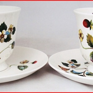 May include: Two white teacups and saucers with a floral pattern featuring green leaves, red strawberries, and yellow flowers. The cups and saucers are made of fine china.
