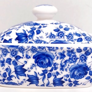 Può includere: Un porta burro in ceramica bianca con un motivo floreale blu e bianco. Il porta burro ha un coperchio con un pomo in cima.