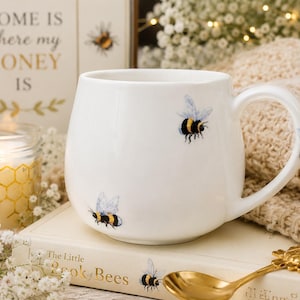 May include: A white ceramic mug featuring a pattern of small, detailed bumblebees with yellow and black stripes and translucent wings. The mug has a rounded body and a curved handle, with the bee illustrations appearing on the exterior surface.