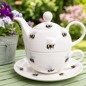 May include: A white ceramic tea set for one, comprising a teapot that stacks onto a matching teacup and saucer. The entire set features a repeating pattern of yellow and black bumblebees with translucent wings across the white surface.