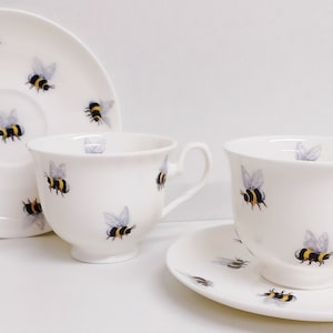 Puede incluir: Dos tazas de té blancas y platillos con un diseño de abeja. Las tazas y los platillos están decorados con abejas negras y amarillas.