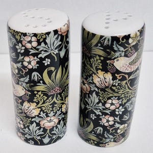 May include: Two ceramic salt and pepper shakers. They feature a black background with a floral and bird pattern in pink, yellow, and green. The design includes flowers, leaves, and small birds. The tops are white with holes.