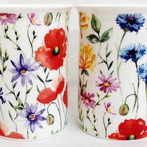 Może przedstawiać: Dwa białe ceramiczne kubki z kwiatowymi wzorami. Jeden kubek przedstawia czerwone maki i fioletowe kwiaty polne, a drugi kubek przedstawia niebieskie chabry i żółte kwiaty polne.