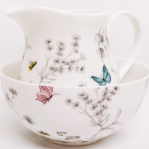 May include: A white ceramic creamer and sugar bowl set with a floral pattern featuring butterflies and bees. The set has a delicate, hand-painted design.