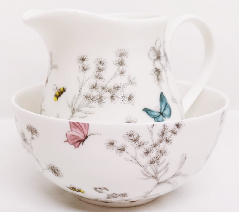 May include: A white ceramic creamer and sugar bowl set with a floral pattern featuring butterflies and bees. The set has a delicate, hand-painted design.