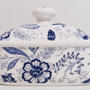 Op de afbeelding: Een witte keramische botervloot met een blauw en wit bloemenpatroon. De vloot heeft een deksel met een knop bovenop.