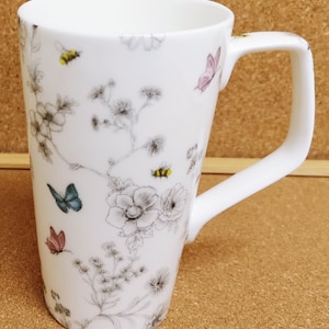 Può includere: Tazza in ceramica bianca con una forma alta e slanciata e un manico grande. La tazza presenta un motivo floreale con farfalle e api in tonalità di rosa, blu, giallo e grigio.