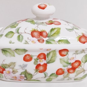 Op de afbeelding: Een witte keramische botervloot met een deksel. De vloot en het deksel zijn versierd met een patroon van rode aardbeien, groene bladeren en roze bloemen.