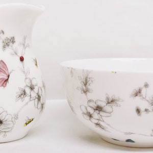 May include: A white ceramic creamer and sugar bowl set with a floral design featuring butterflies and bees. The design is in shades of gray, pink, and yellow.