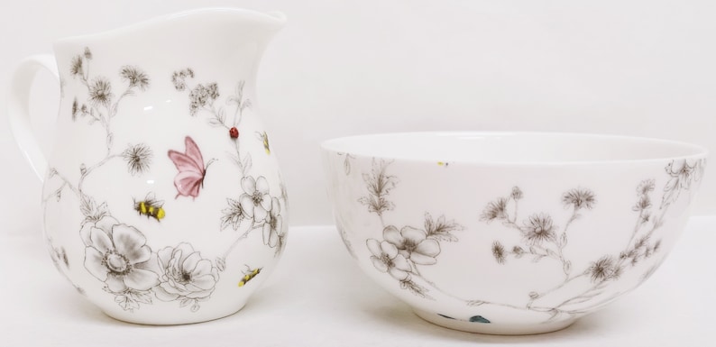 May include: A white ceramic creamer and sugar bowl set with a floral design featuring butterflies and bees. The design is in shades of gray, pink, and yellow.