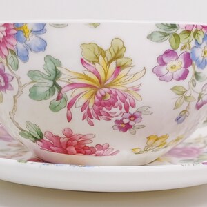 May include: A white teacup and saucer set with a floral pattern. The pattern features pink, yellow, blue, and green flowers on a white background.