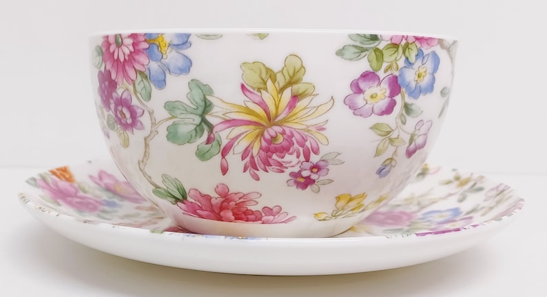 May include: A white teacup and saucer set with a floral pattern. The pattern features pink, yellow, blue, and green flowers on a white background.