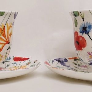 Op de afbeelding: Twee witte theekopjes en schoteltjes met een bloemenpatroon. De kopjes en schoteltjes hebben een aquarel-achtig ontwerp met verschillende bloemen in tinten roze, blauw, geel en rood.