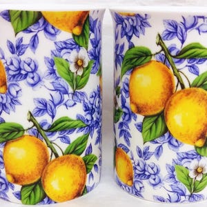 May include: Two white ceramic mugs featuring a blue floral pattern and yellow lemons. Each mug has a curved handle and a design of lemons with green leaves and white flowers. Perfect for tea or coffee.