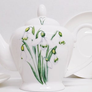 May include: White tea set with a floral design of white snowdrop flowers with green leaves. The set includes a teapot, two teacups, and two saucers.