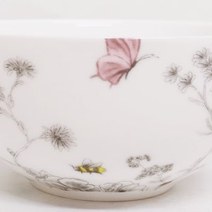 May include: White ceramic bowl with a floral design featuring a pink butterfly and a bee. The bowl has a delicate, hand-painted look.