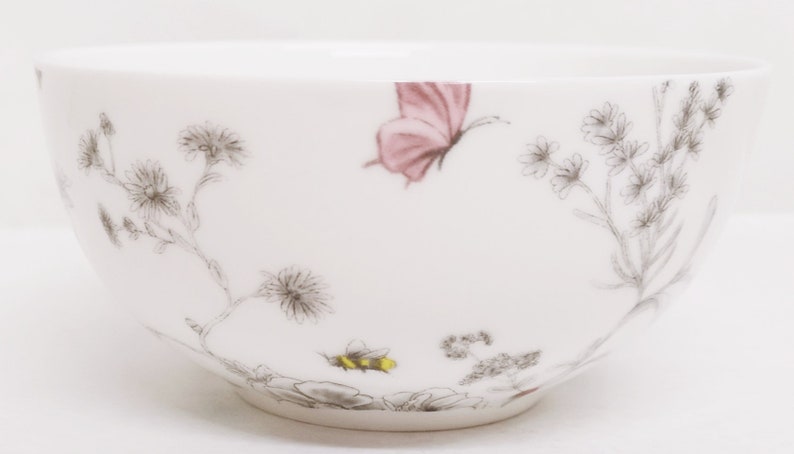 May include: White ceramic bowl with a floral design featuring a pink butterfly and a bee. The bowl has a delicate, hand-painted look.