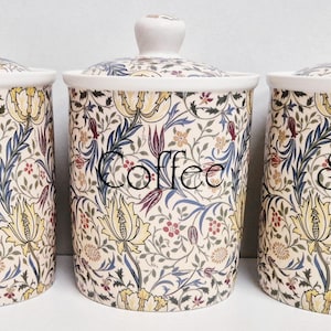 Può includere: Tre barattoli in ceramica con coperchi, etichettati "Tea", "Coffee" e "Sugar". Ogni barattolo presenta uno sfondo color crema con un motivo floreale giallo, blu e rosso. I coperchi hanno un piccolo pomello sopra.