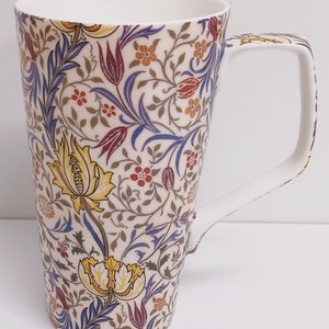 Op de afbeelding: Een hoge witte keramische mok met een bloemenpatroon in tinten geel, blauw, rood en bruin. De mok heeft een groot handvat.
