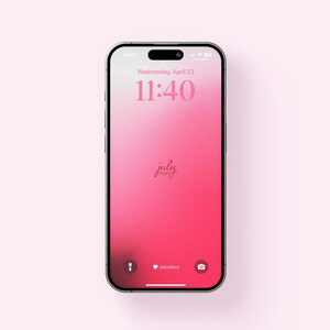 以下が含まれることがあります： ピンクのグラデーションの壁紙が表示されたスマートフォン。ロック画面には日付、時刻、そして「july」の文字が表示されています。スマートフォンは薄いピンク色の背景にあります。