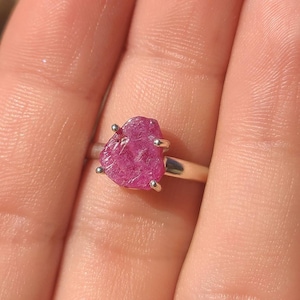 May include: A silver ring featuring a raw, pink ruby gemstone. The ruby is set in a prong setting and has a rough, natural appearance.