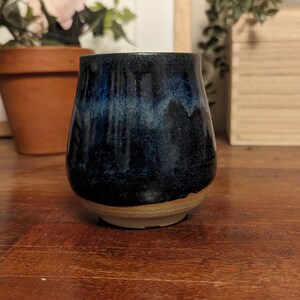 May include: A dark blue ceramic cup with a light blue glaze and a brown rim. The cup is sitting on a wooden table.