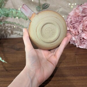 以下が含まれることがあります： 赤い取っ手が付いた、薄い茶色の小さな陶器のマグカップ。マグカップの底には円形の模様があり、中央にシンボルがあります。