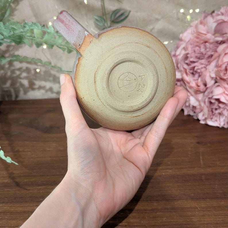 以下が含まれることがあります： 赤い取っ手が付いた、薄い茶色の小さな陶器のマグカップ。マグカップの底には円形の模様があり、中央にシンボルがあります。