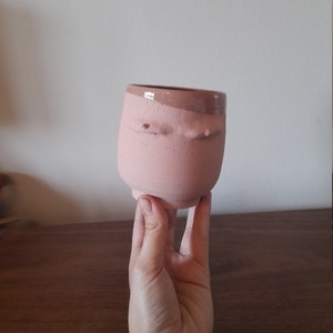 May include: A pink ceramic cup with a textured surface and a brown rim. The cup has a unique design with two raised bumps on the front.