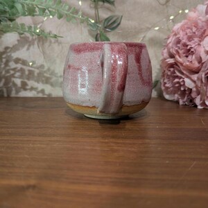以下が含まれることがあります： ピンクと白の釉薬をかけた手作りセラミックマグカップ。マグカップは丸みを帯びたボディとハンドルが付いています。