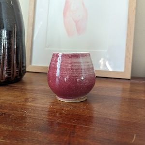 May include: A small, round, ceramic cup with a deep red glaze and a white rim. The cup has a textured surface and sits on a light-coloured base. A dark brown vase is to the left, and a framed drawing is in the background.