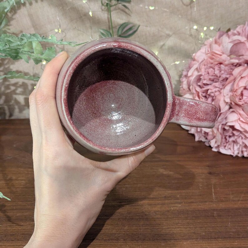以下が含まれることがあります： 白とピンクの釉薬をかけたセラミック製のマグカップ。マグカップは丸みを帯びた形をしていて、持ち手があります。