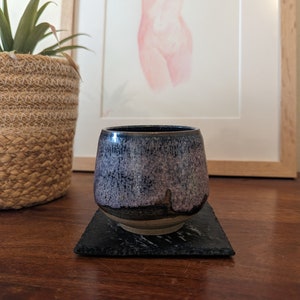 May include: A dark blue and purple ceramic cup with a speckled glaze sits on a black slate coaster. The cup is slightly wider at the top than the bottom.