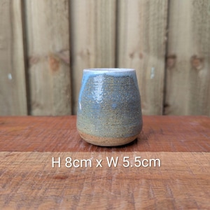 Könnte beinhalten: Ein handgefertigter Keramikbecher mit blau-beiger Glasur. Der kleine, abgerundete Becher ist 8 cm hoch und 5,5 cm breit. Der Boden ist unglasiert und zeigt die natürliche Tonfarbe. Der Innenraum ist weiß.