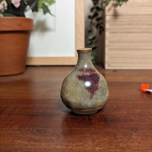May include: A small, round, grey ceramic vase with a red and brown glaze. The vase is sitting on a wooden table.