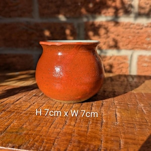 May include: A small, round, orange ceramic pot with a speckled texture. The pot has a white interior and a slightly ruffled rim. The dimensions are approximately 2.75 inches high and 2.75 inches wide. The pot sits on a wooden surface.
