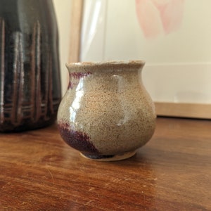 May include: A small, round, ceramic vase with a speckled beige glaze and a dark burgundy band around the bottom.