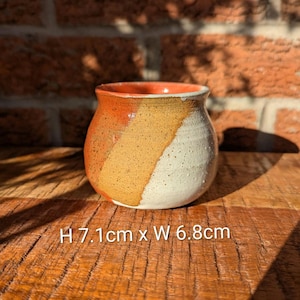 May include: A handmade ceramic pot with a rounded shape. The pot features a colour scheme of orange, tan, and white. The dimensions are approximately 7.1 cm high and 6.8 cm wide. The pot is sitting on a wooden surface.