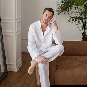 May include: A person wearing a white linen pajama set, including a button-down shirt and pants. The person is seated on a brown sofa in a well-lit room with a palm plant. The floor is hardwood with a patterned rug.