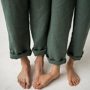 May include: Close-up of three pairs of legs wearing dark green linen pants with rolled-up cuffs. The bare feet of the wearers are visible against a white background. The pants have a relaxed fit and a slightly wrinkled texture.