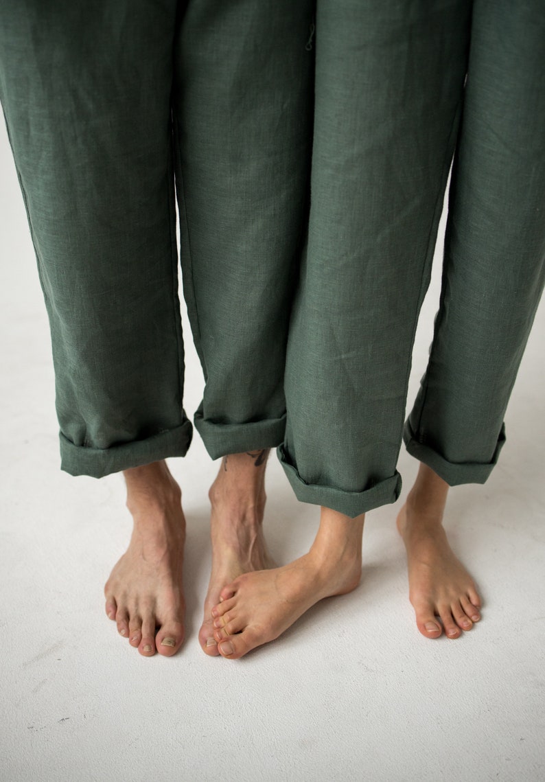 May include: Close-up of three pairs of legs wearing dark green linen pants with rolled-up cuffs. The bare feet of the wearers are visible against a white background. The pants have a relaxed fit and a slightly wrinkled texture.